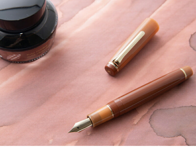 A brown fountain pen with a gold nib and trim, shown with its cap removed on a pinkish textured surface. Part of a dark ink bottle is visible in the upper left.